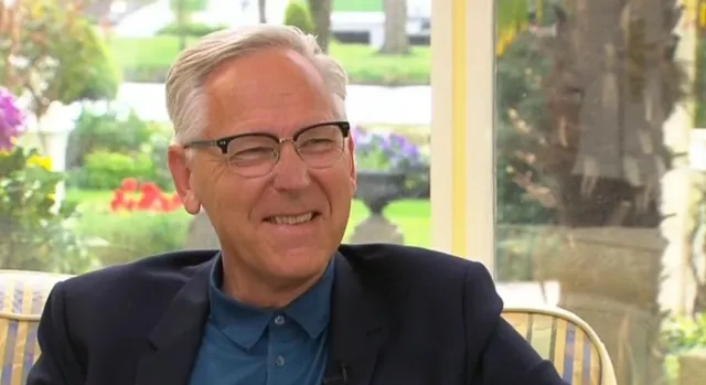 Jan Pool zit op een stoel in een serre met op de achtergrond een tuin.